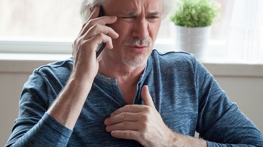 Man with squinted face covers the chest with his hand while on the phone. He is noticeably in the pain or experiencing heartache.